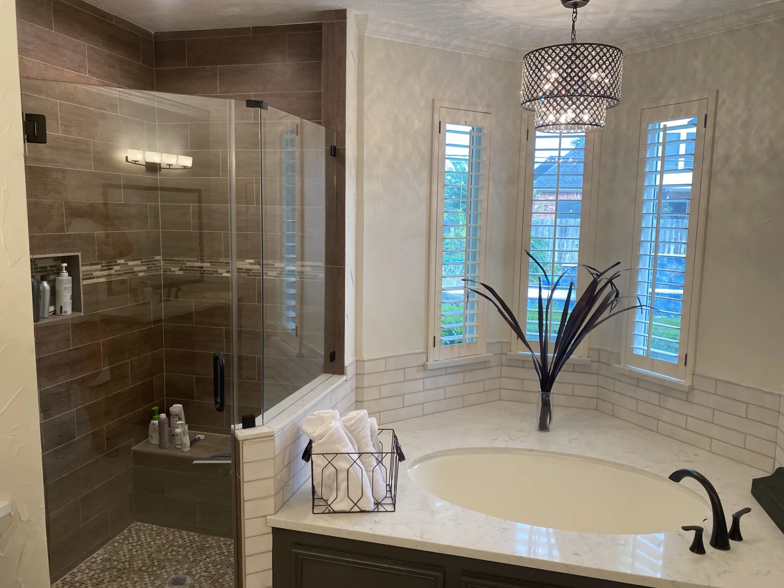 Renovated master bathroom featuring a frameless glass shower with wood-look tile, white subway tile soaking tub surround, and a crystal drum chandelier