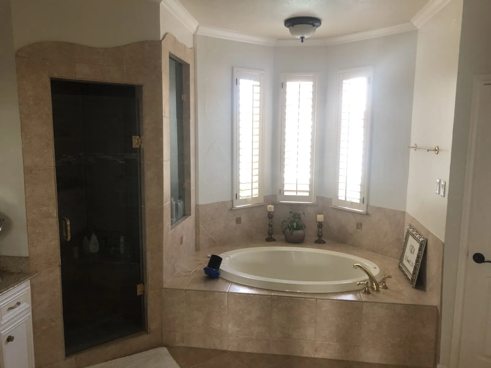 Dated master bathroom with beige tile surround, oval soaking tub, dark glass-door shower enclosure, and plantation shutters