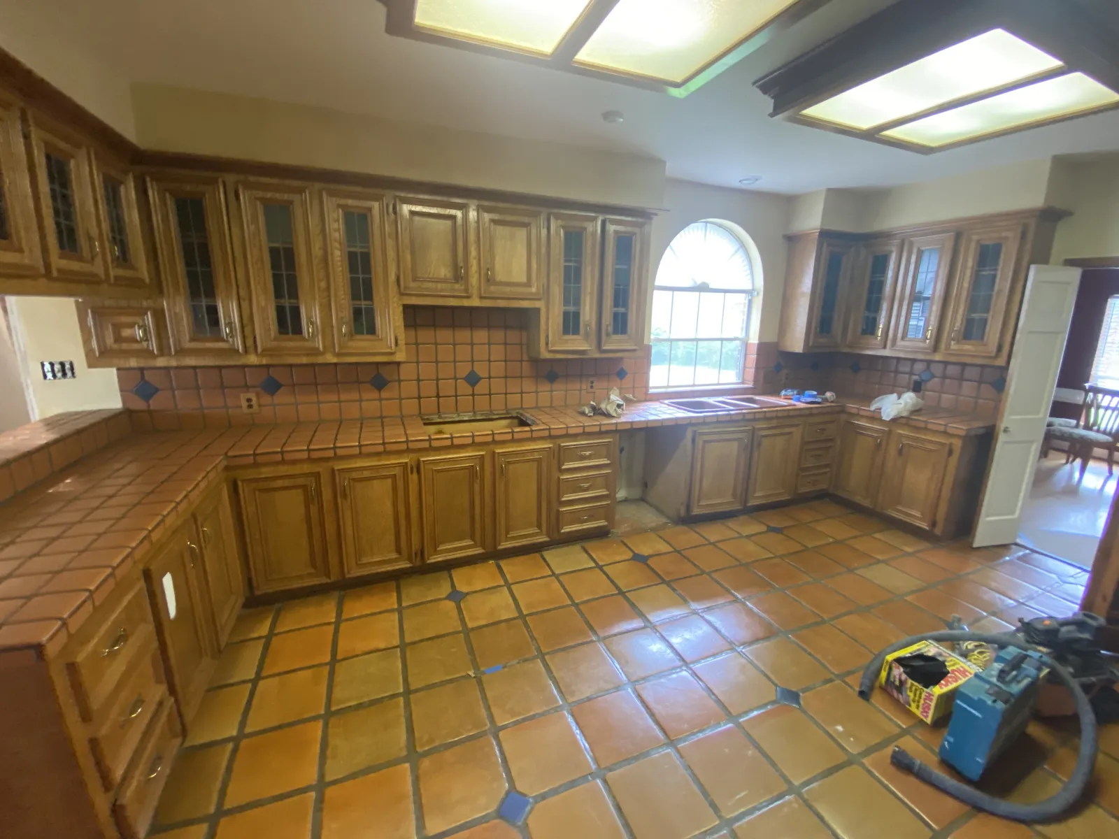 Wide-angle view of a dated kitchen with oak raised-panel cabinets, Saltillo tile countertops and floors, fluorescent box lighting, and an arched window