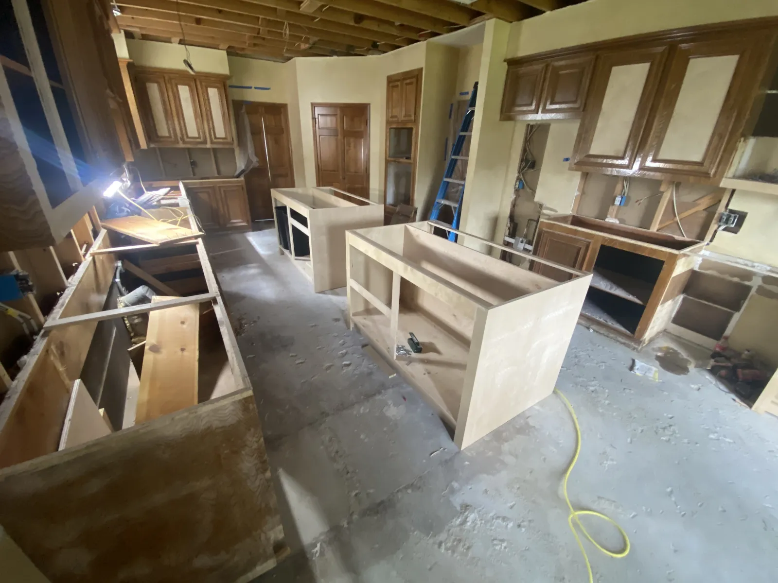 Kitchen mid-renovation with bare concrete floors, new unfinished cabinet boxes being installed, exposed ceiling framing, and construction tools