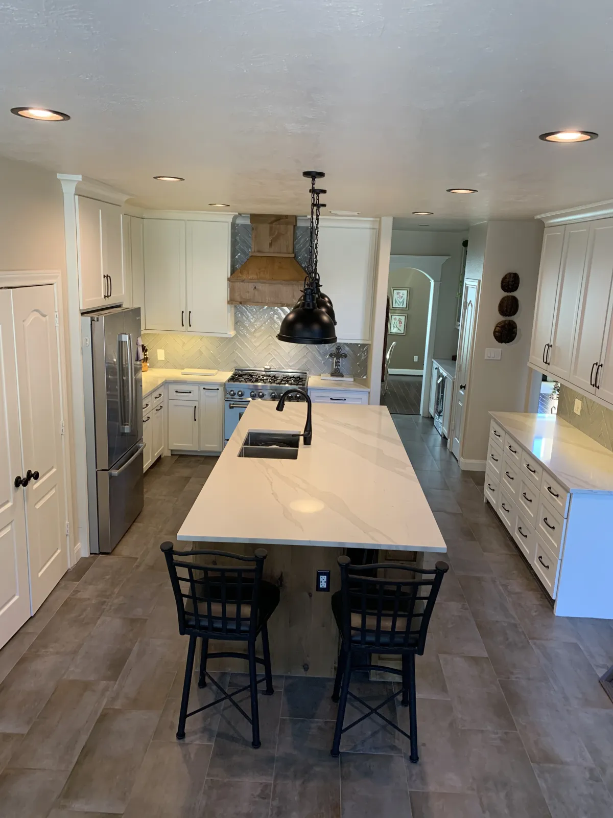 A beautifully renovated kitchen featuring white shaker cabinets, a large waterfall-edge quartz island with bar seating, herringbone tile backsplash, and wood-look tile flooring