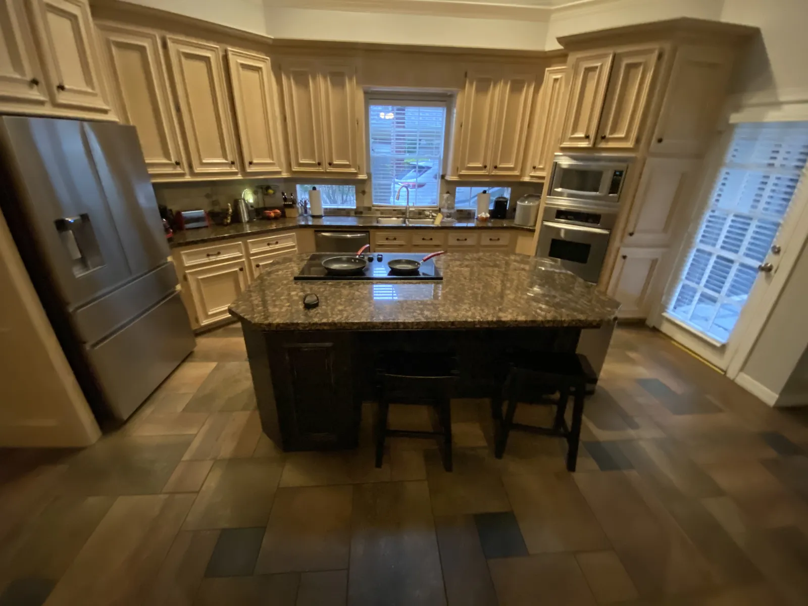 A closer view of the dated kitchen showing cream glazed cabinets, granite island with cooktop, and stainless steel appliances