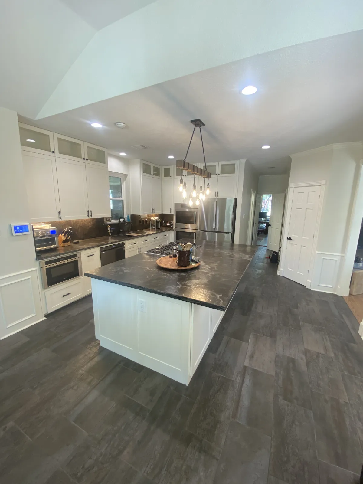 A fully remodeled kitchen with white shaker cabinets, dark marbled quartz countertops, a rustic wood-beam chandelier with Edison bulbs, and dark porcelain tile flooring