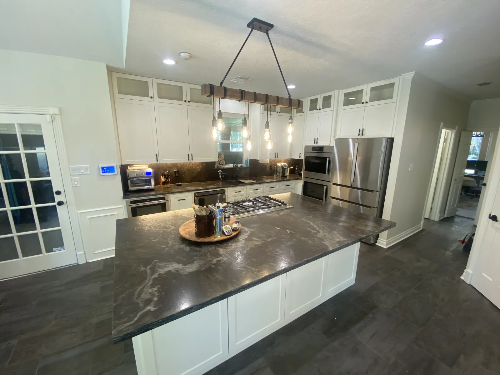 The completed white and dark quartz kitchen from the island perspective, showing the expansive countertop with gas cooktop and wood-beam Edison bulb chandelier