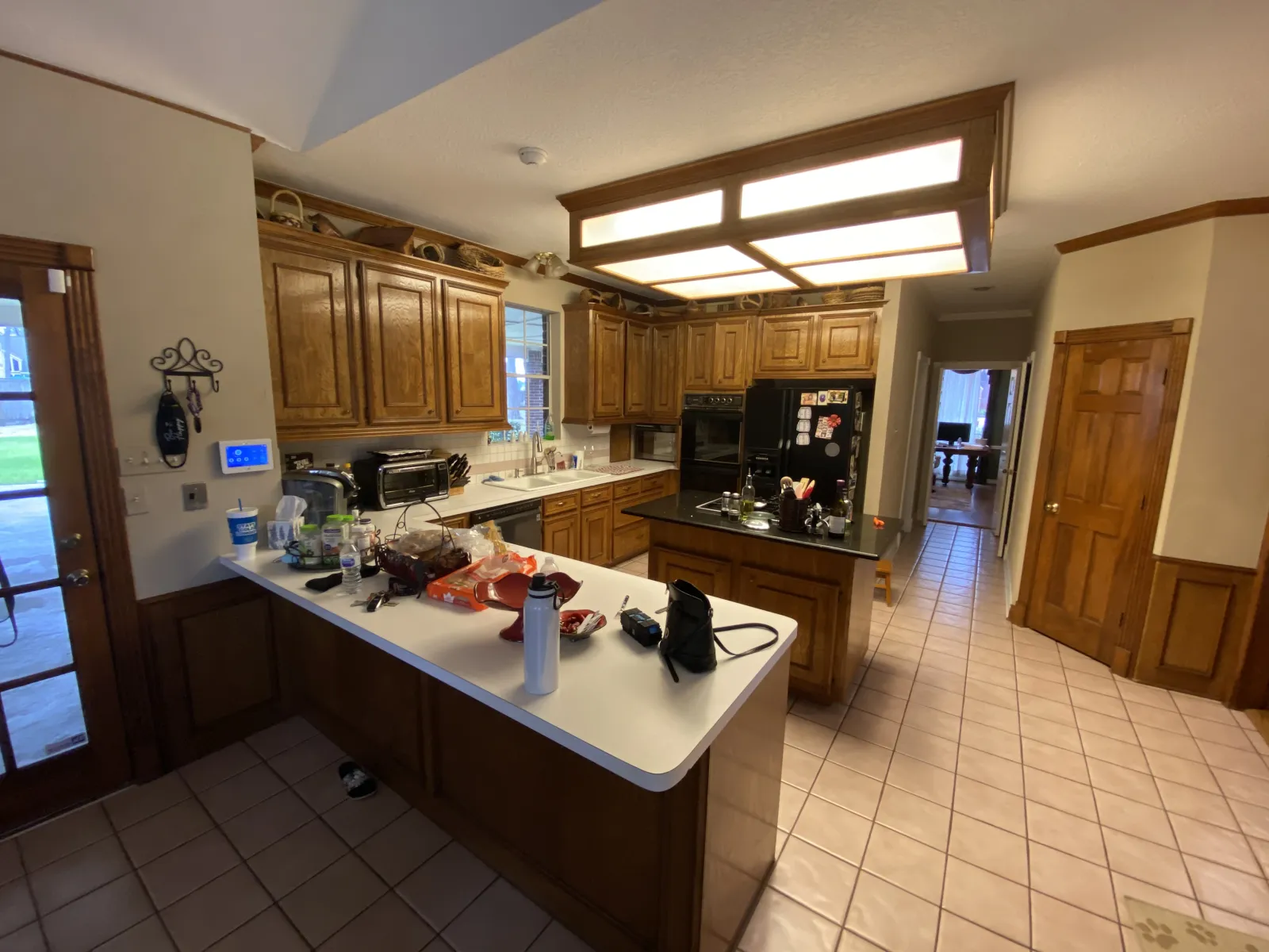 A spacious but outdated kitchen with honey oak cabinets, white laminate countertops, fluorescent box lighting, and beige ceramic tile flooring