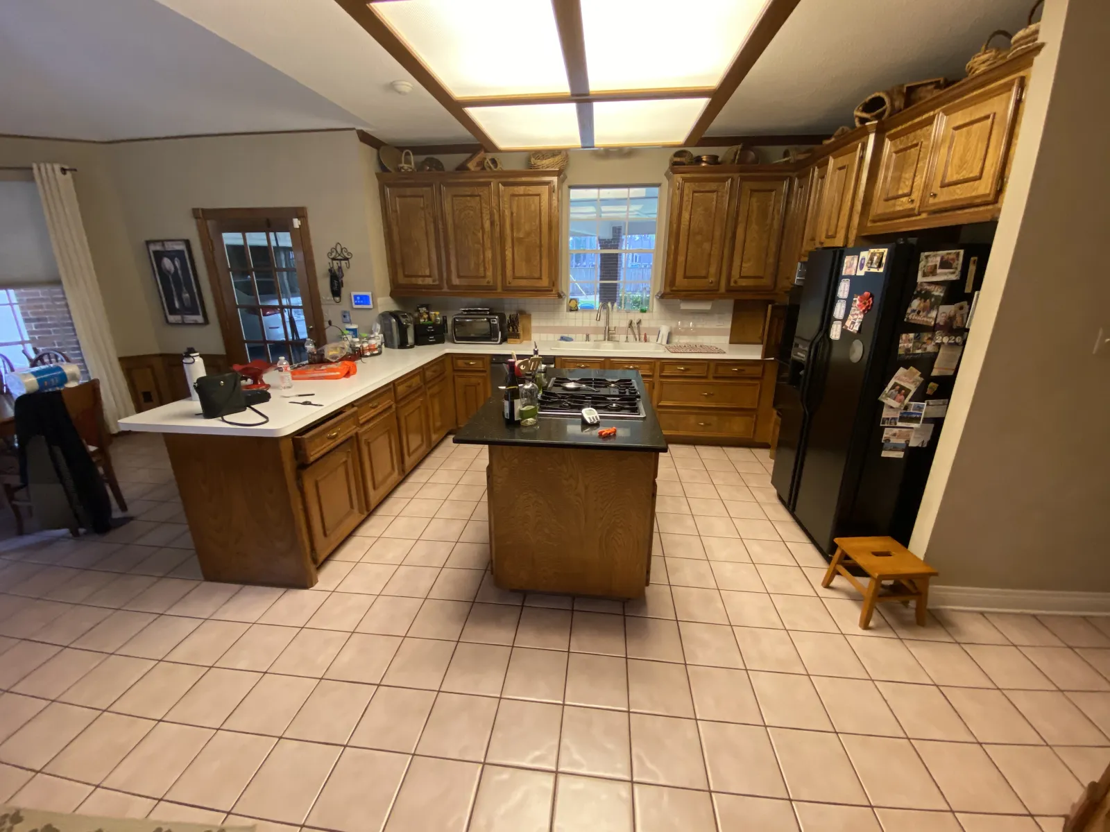 An alternate angle of the dated oak kitchen showing the small cooktop island, fluorescent box light, and open layout