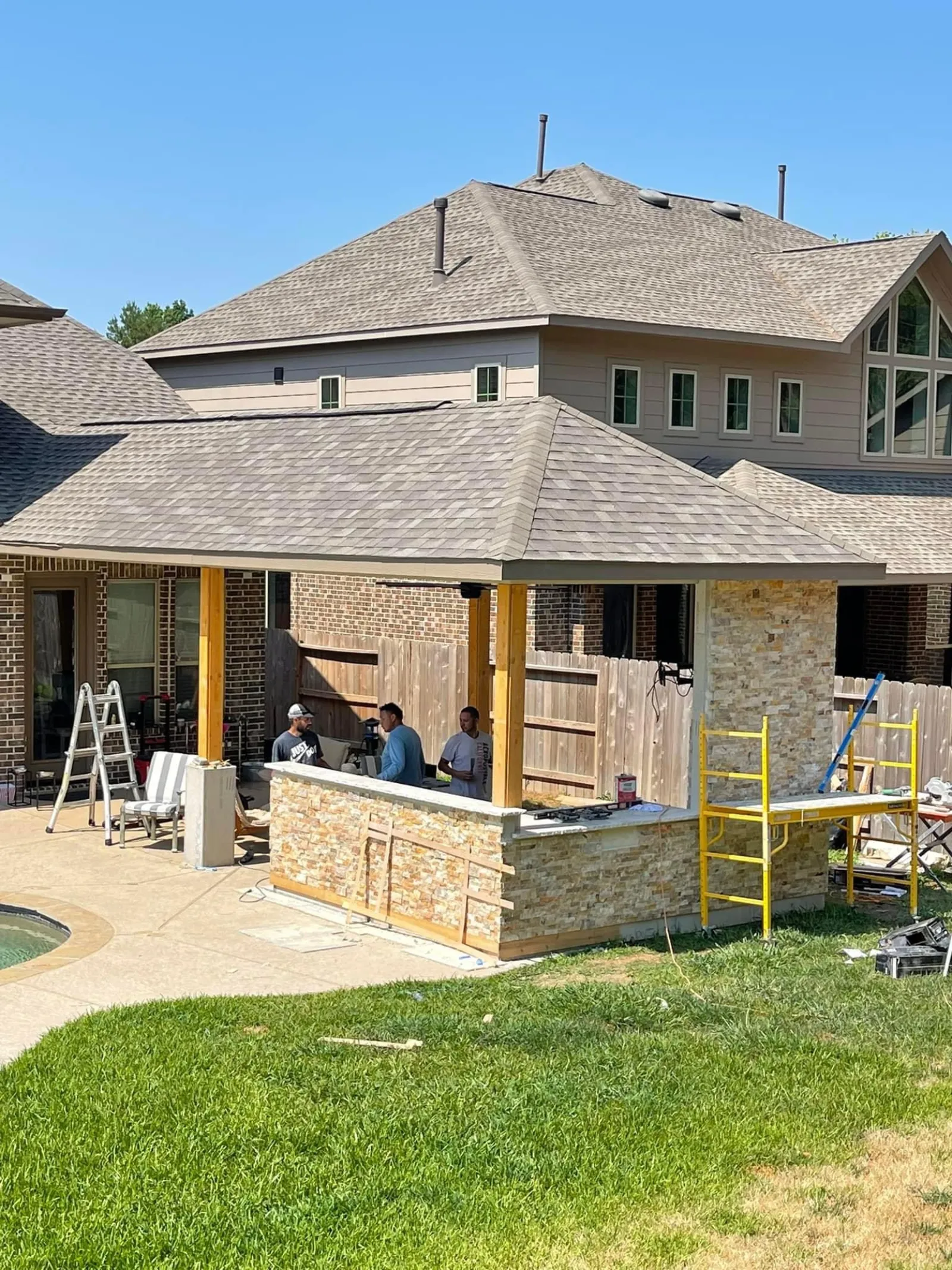 Crew members applying stone veneer to an outdoor kitchen bar under a newly roofed covered patio