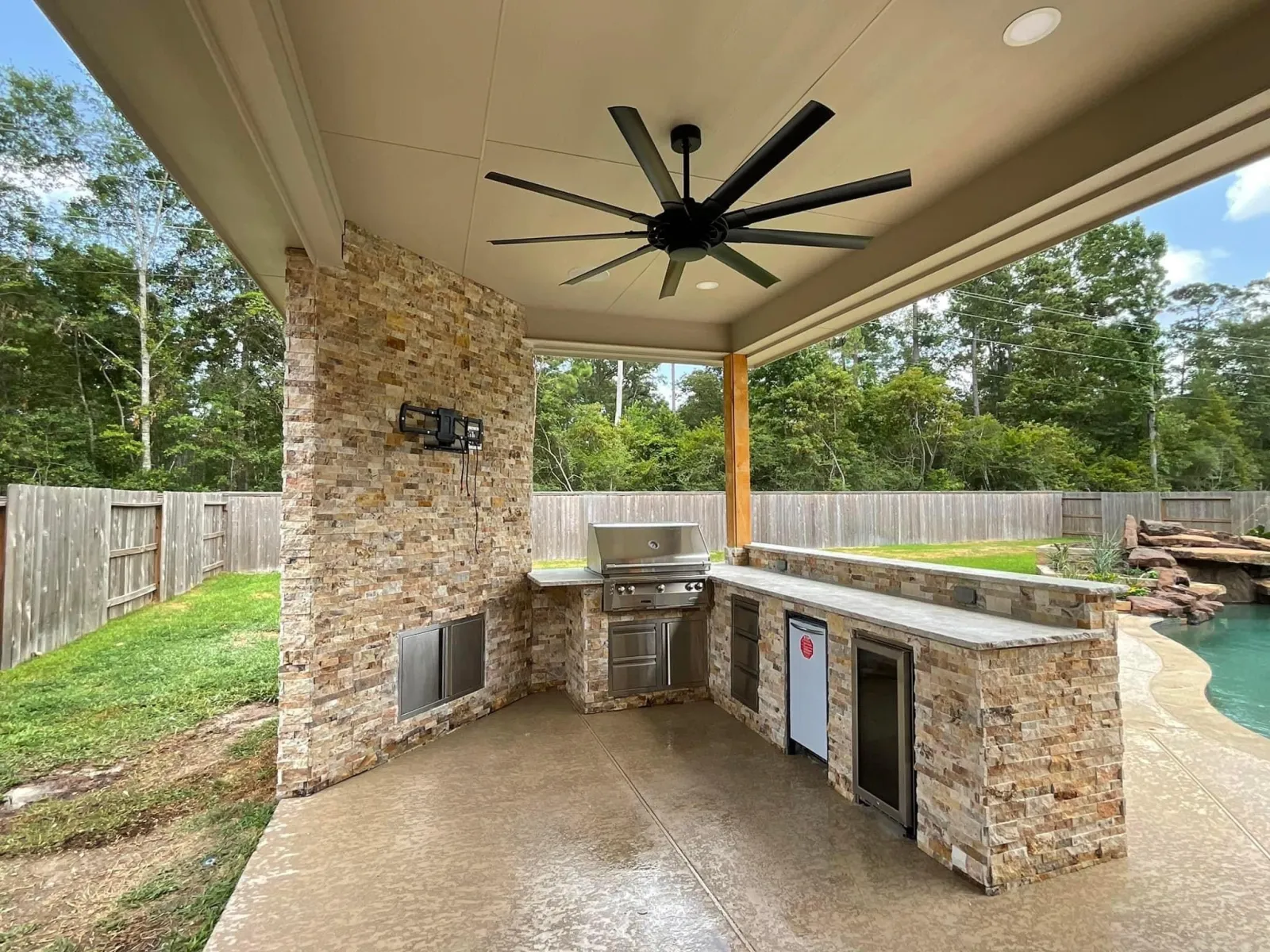 Interior view of a finished outdoor kitchen under a covered patio with stone veneer, built-in grill, and refrigerator