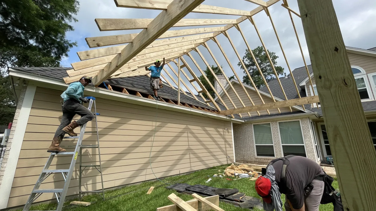 Workers installing roof rafters on a large gabled covered patio frame attached to the back of a two-story home